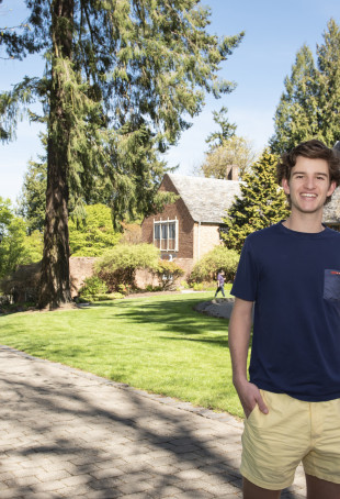 Bennett smiling outside by the Manor House on the undergraduate campus. He is wearing a navy tshirt and yellow shorts.